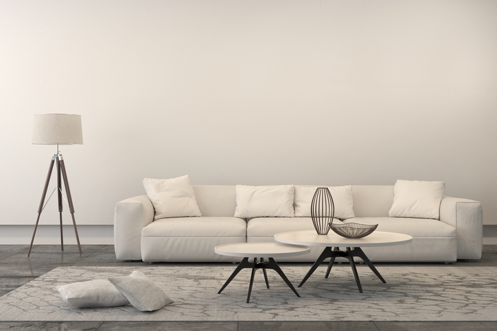White sofa with set of coffee tables, a floor lamp and pillows. Interior is white, brightly lit with ornate carpet. Concrete industrial floor and a white wall behing. Copy space. 3D render